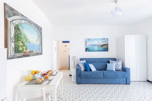 A bright living room with a blue sofa, a dining table with breakfast, and a large painting of a coastal scene. - SEA FRONT TERRACE APARTMENT MARINA GRANDE SORRENTO (Sorrento)