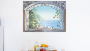A breakfast setup with a painting of a coastal scene, a pitcher of orange juice, a bowl of fruit, and pastries on a table. - SEA FRONT TERRACE APARTMENT MARINA GRANDE SORRENTO (Sorrento)