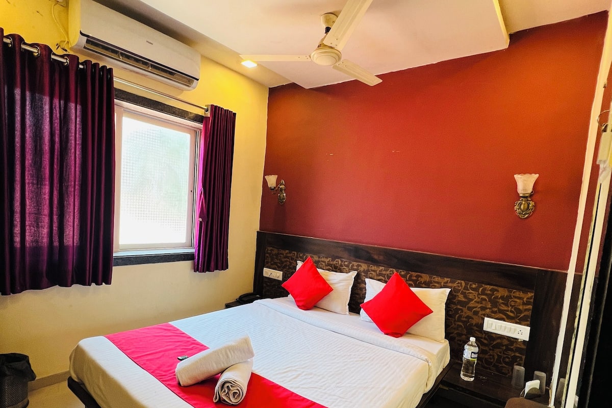 A hotel room with a large bed, red and white bedding, a ceiling fan, a window with purple curtains, and a wall-mounted nightstand with a bottle of water.