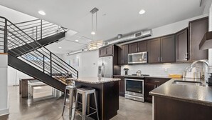 Una cocina moderna con armarios de madera oscura, electrodomésticos de acero inoxidable, una isla central con taburetes y una escalera que sube a un nivel superior.