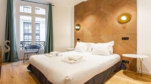 Ein Hotelzimmer mit einem großen Bett, einem kleinen runden Tisch, einem Sessel und Blick auf die Stadt durch ein Fenster.