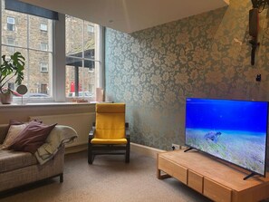 Ein Wohnzimmer mit einer Couch, einem gelben Sessel, einer Holz-TV-Konsole und einem großen Fernseher, der eine Unterwasserlandschaft zeigt.