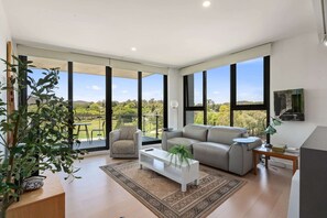 Una sala de estar moderna con un sofá gris, una mesa de centro blanca y un ventanal con vistas a un jardín y colinas.