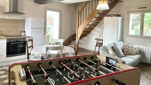 A cozy living room with a foosball table, a dining area with a table and chairs, a kitchen with a stove and refrigerator, and a staircase leading to another level. - La Maison Agoméa (Saint-Georges-sur-Cher)
