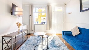 Ein modernes Wohnzimmer mit einem blauen Sofa, einem an der Wand montierten Fernseher, einem kleinen runden Tisch und einem großen Fenster mit Vorhängen.