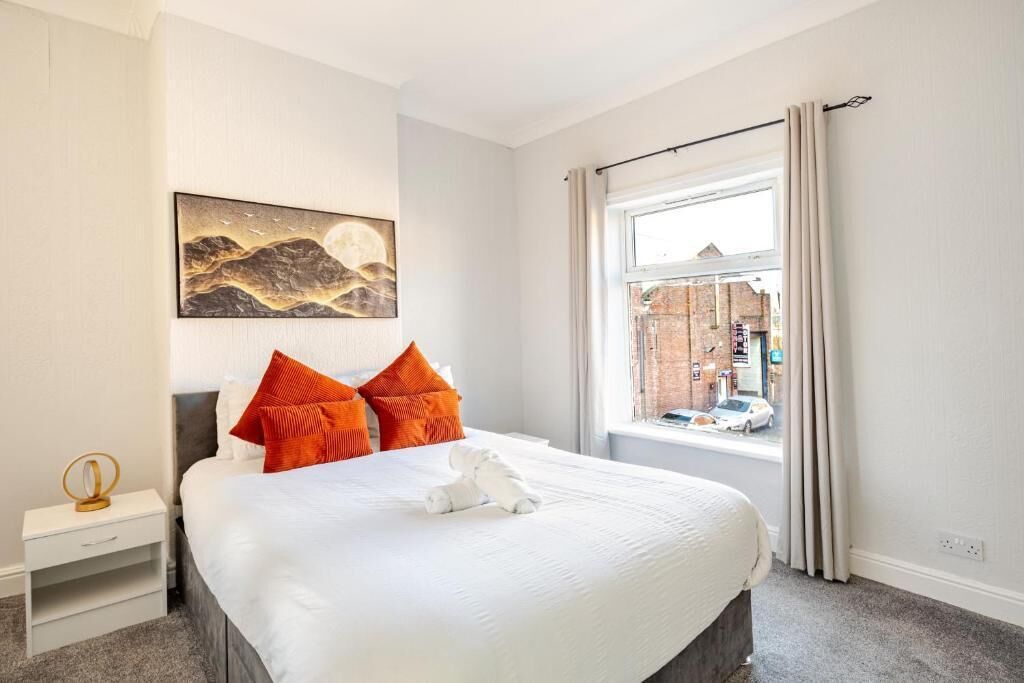 Ein ordentlich gemachtes Bett mit weißer Bettwäsche und orangen Kissen, ein Nachttisch mit Lampe sowie ein großes Fenster mit Blick auf eine Straße und Gebäude.