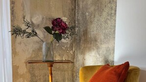 Un fauteuil jaune avec un coussin rouge, une petite table en bois sur laquelle est posé un vase de fleurs, et un mur en béton.