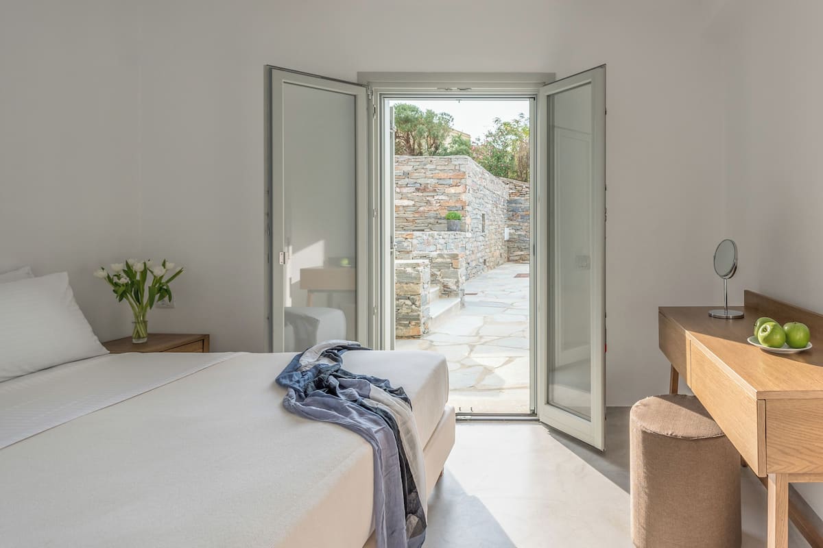 A bedroom with a bed, a bedside table with a vase of flowers, a wooden desk with a mirror and apples, and a stone wall outside.