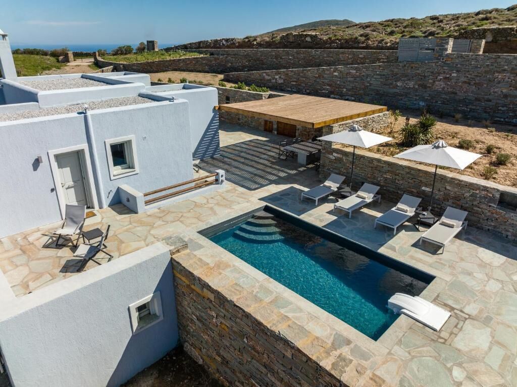 Un área de piscina en la azotea con sillones de descanso y sombrillas, rodeada de muros de piedra y vistas al paisaje.