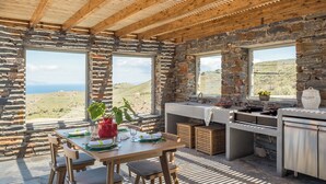 A rustic stone interior with a wooden table set for dining, a sink, and a view of the landscape. - NEVERMIND VACATIONS VILLAS (Kea)