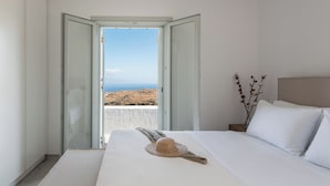 A bedroom with a bed, a view of the sea, and a window with an open door. - NEVERMIND VACATIONS VILLAS (Kea)