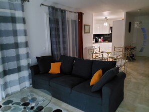 Un salon avec un canapé foncé, des coussins orange, une table basse en verre et un coin repas comprenant une table et des chaises.