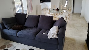 Una sala de estar moderna con un sofá azul oscuro, una mesa de comedor con superficie de vidrio y sillas, y una cocina al fondo.