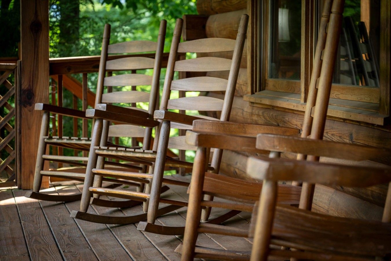 A wooden rocking chair on a porch with a wooden railing and a window.