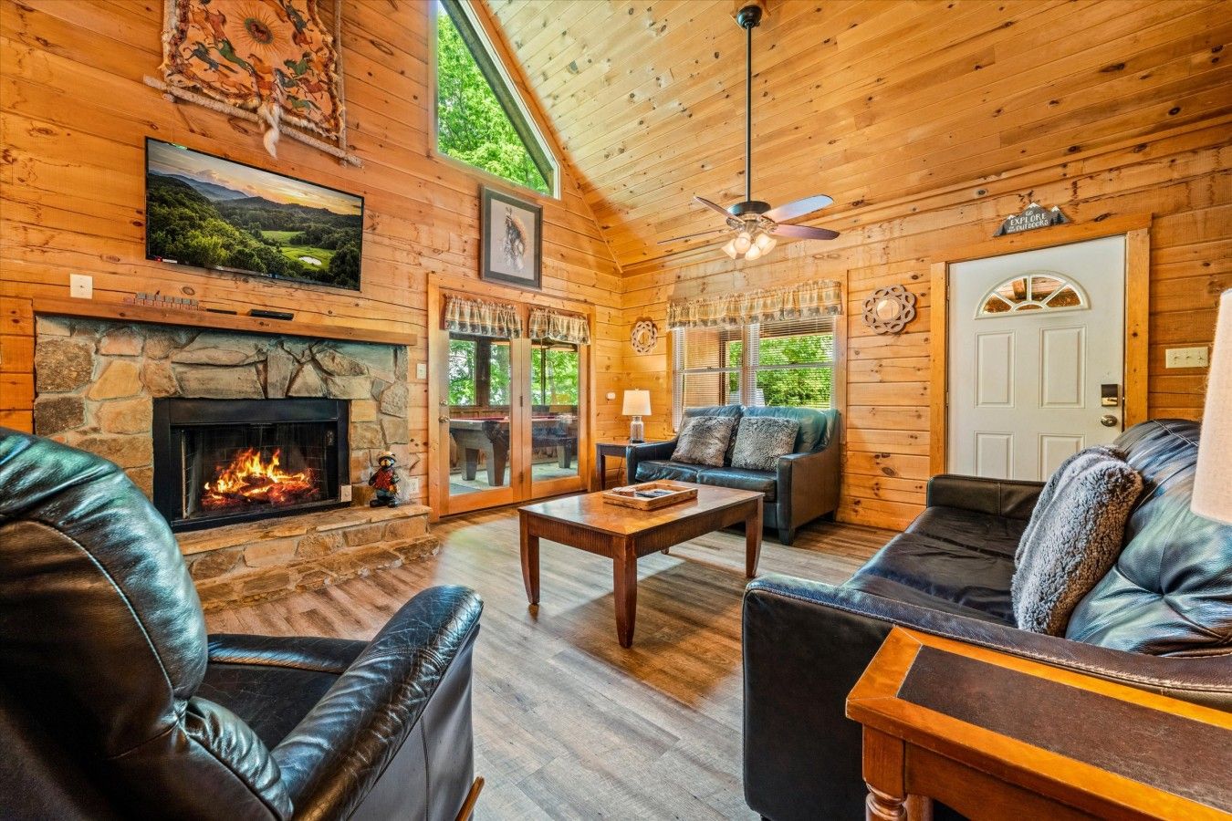 L’intérieur chaleureux d’un chalet avec une cheminée en pierre, des meubles en cuir et une grande fenêtre donnant sur un paysage verdoyant.