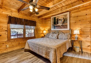 A wooden cabin bedroom with a bed, a ceiling fan, a window with blinds, a framed picture on the wall, and a nightstand with a lamp. - Peaceful 2BR w/Hot Tub*Pool Table*Fireplace*Arcade (Sevierville)