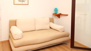Un divano beige con struttura in legno, una piccola lampada color petrolio su uno scaffale di legno e un quadro appeso al muro.