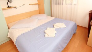 Un letto rifatto con copriletto blu e bianco, due asciugamani piegati e una scrivania con lampada in una stanza con pavimento in legno e pareti di colore chiaro.