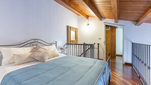 Quarto com cama, vigas de madeira no teto, escada e porta que dá para outro cômodo.