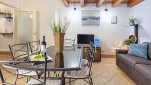 Uma área de refeições com mesa de vidro, cadeiras de metal, um vaso com plantas, uma televisão e um sofá.