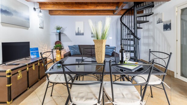 Uma área de refeições com mesa de vidro, cadeiras de metal, um cesto trançado como peça central e uma escada.