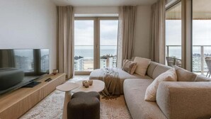 A modern living room with a large flat-screen TV, a sofa, a coffee table, and a view of the city through large windows. - Nora - Elegant Seaview Escape With Balcony (Ostend)