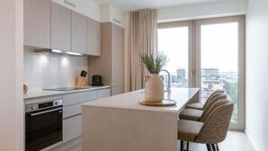 A modern kitchen with a central island, a dining area with four chairs, and a large window with a city view. - Nora - Elegant Seaview Escape With Balcony (Ostend)