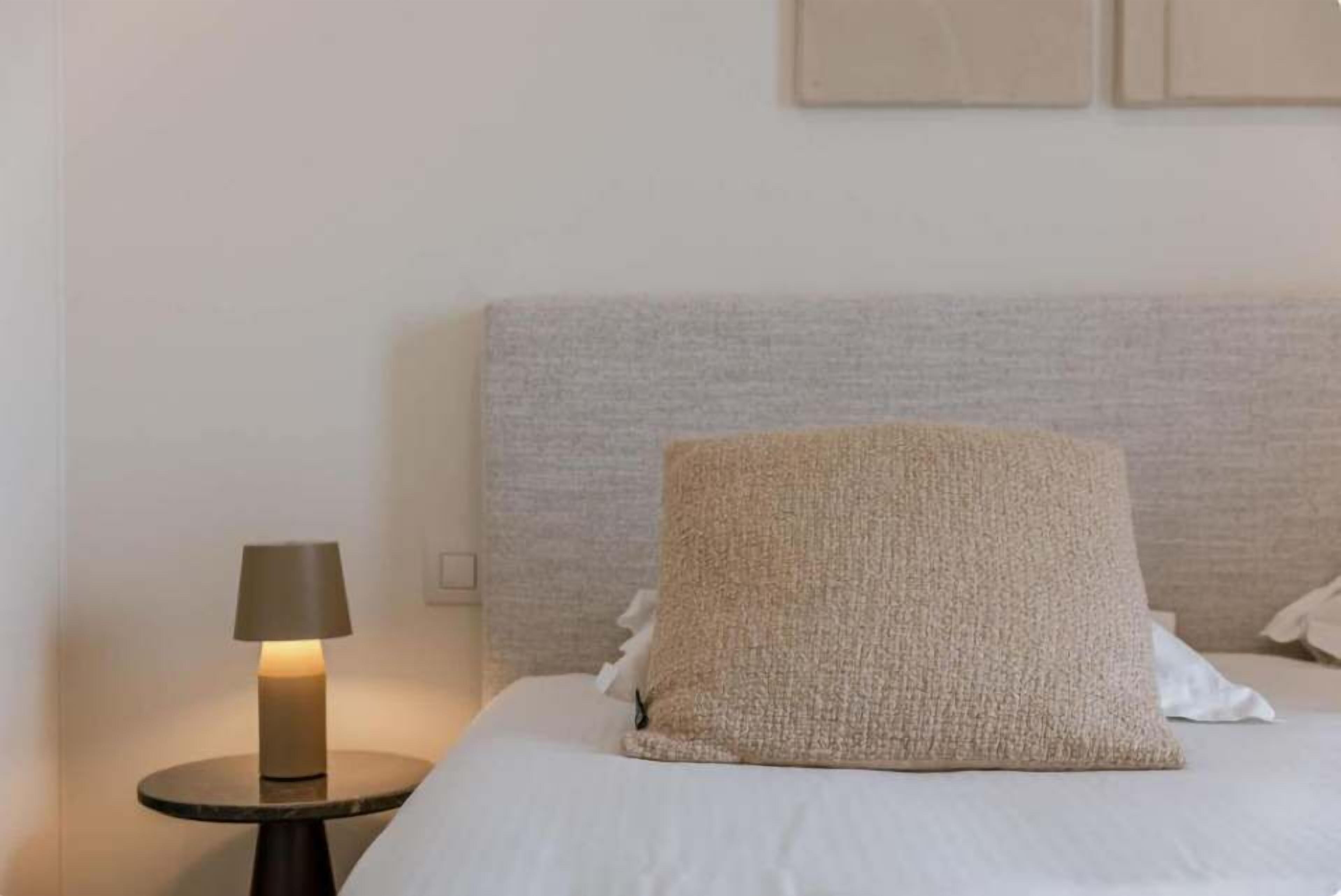 Een net opgemaakt bed met een ge текстуреerd kussen en een klein nachtkastje met een lamp.