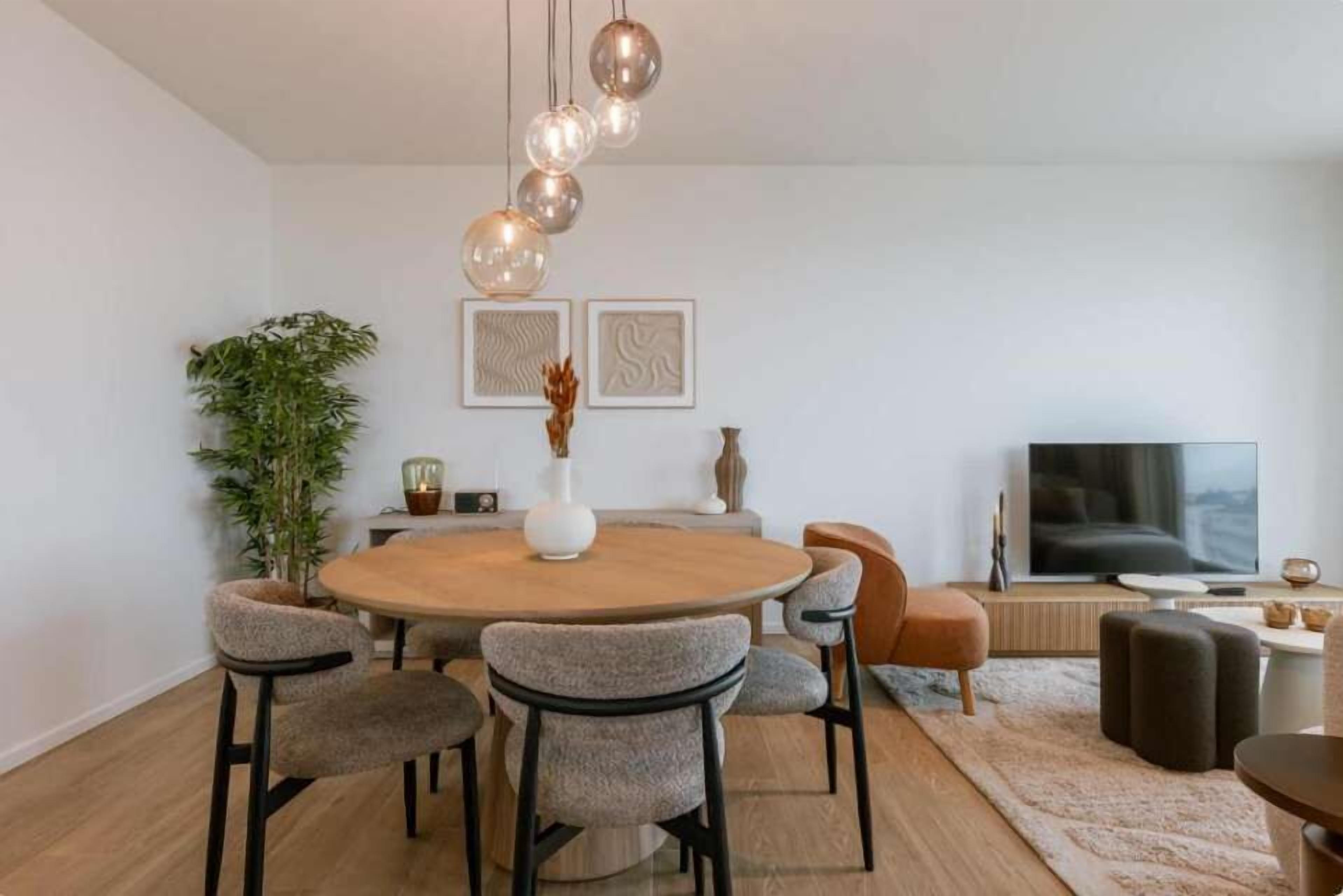 A modern dining area with a round wooden table, six upholstered chairs, a potted plant, and a television on a stand.