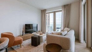 A modern living room with a sofa, ottomans, a TV, and a view of the city through large windows. - Nora - Elegant Seaview Escape With Balcony (Ostend)