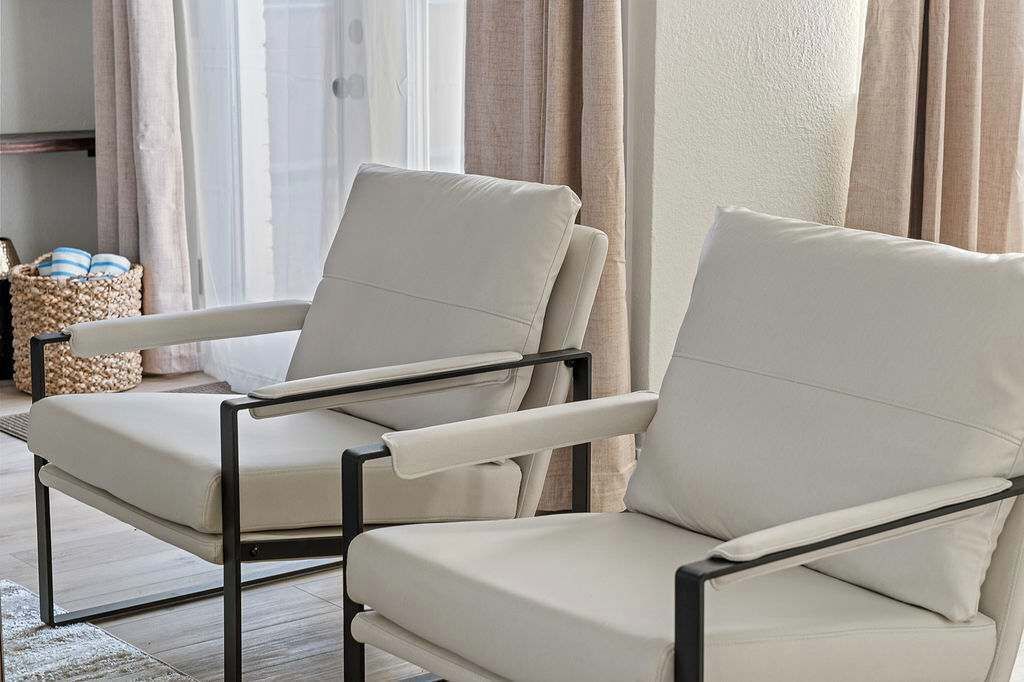 Two beige armchairs with black metal frames, a woven basket, and a window with curtains.