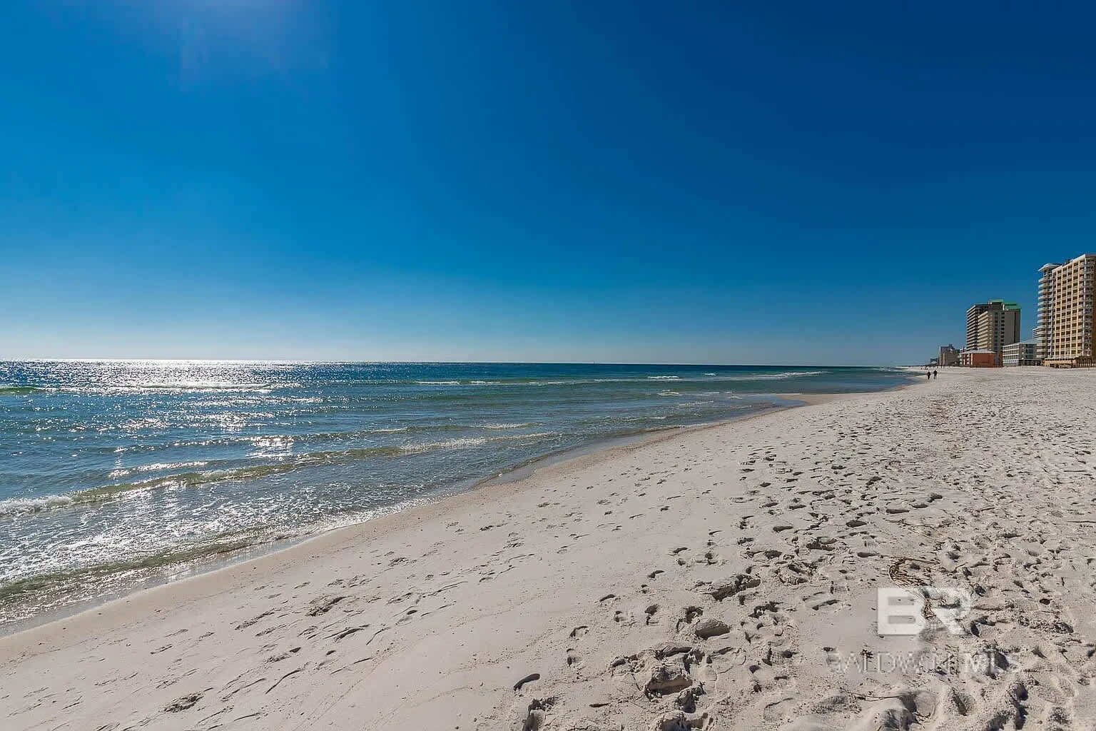 A beach with footprints leading to the water, a clear sky, and buildings in the background.