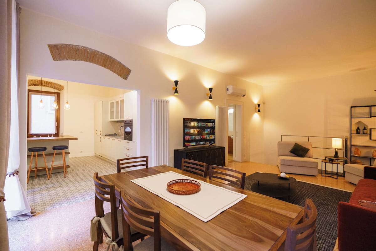 A dining area with a wooden table, chairs, and a central island. There is a kitchen area with cabinets and a refrigerator in the background.