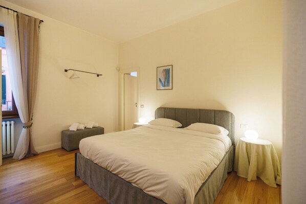 Ein Schlafzimmer mit einem Bett, Nachttischen, einer Bank, einem Heizkörper und einem Fenster mit Vorhängen.