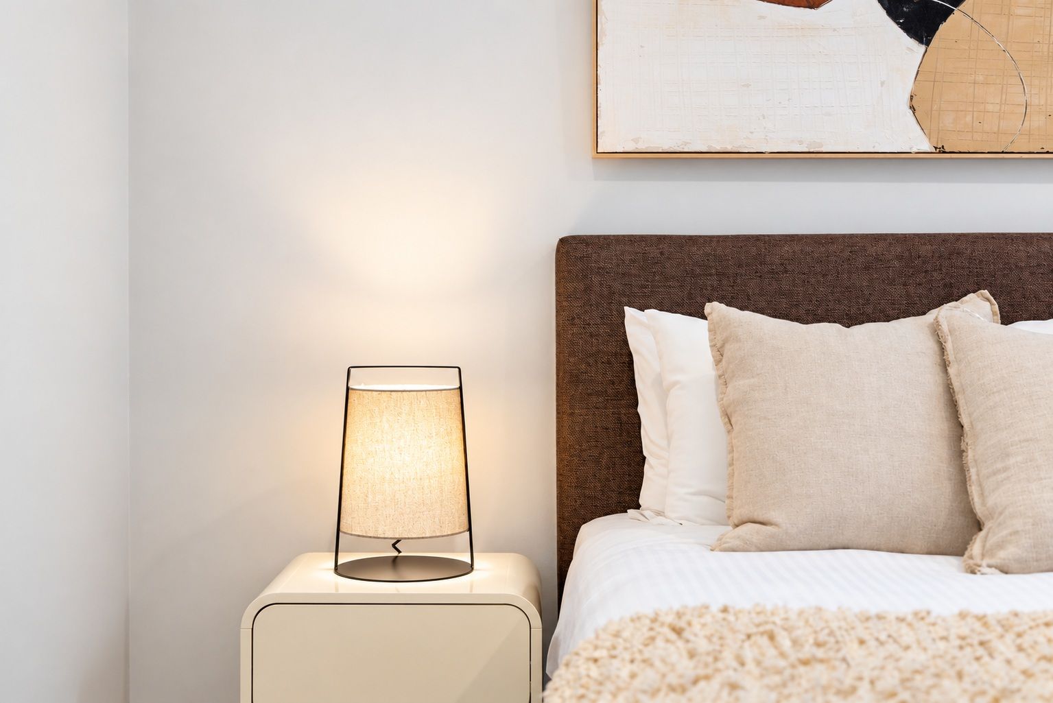 Ein ordentlich bezogenes Bett mit Kopfteil, einer Nachttischlampe und einem an der Wand hängenden abstrakten Gemälde.