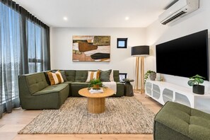 Ein modernes Wohnzimmer mit einem Ecksofa, einem Couchtisch, einem Flachbildfernseher und einem großen Fenster mit Vorhängen.