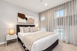 Ein modernes Schlafzimmer mit einem großen Bett, einem Kopfteil, einem Nachttisch mit Lampe und einem Fenster mit Vorhängen.