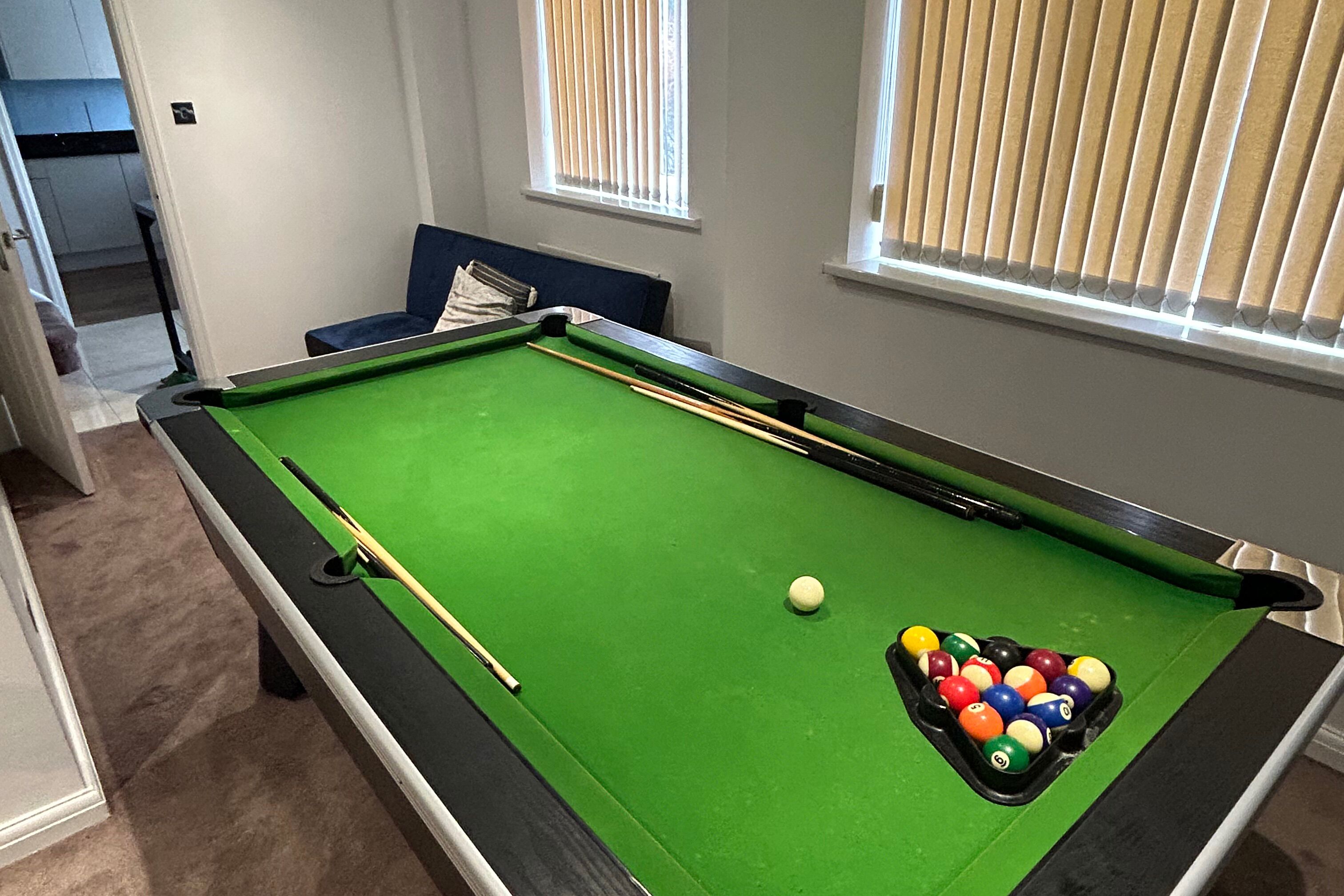 A pool table with balls and cues, a blue chair, and a window with blinds.
