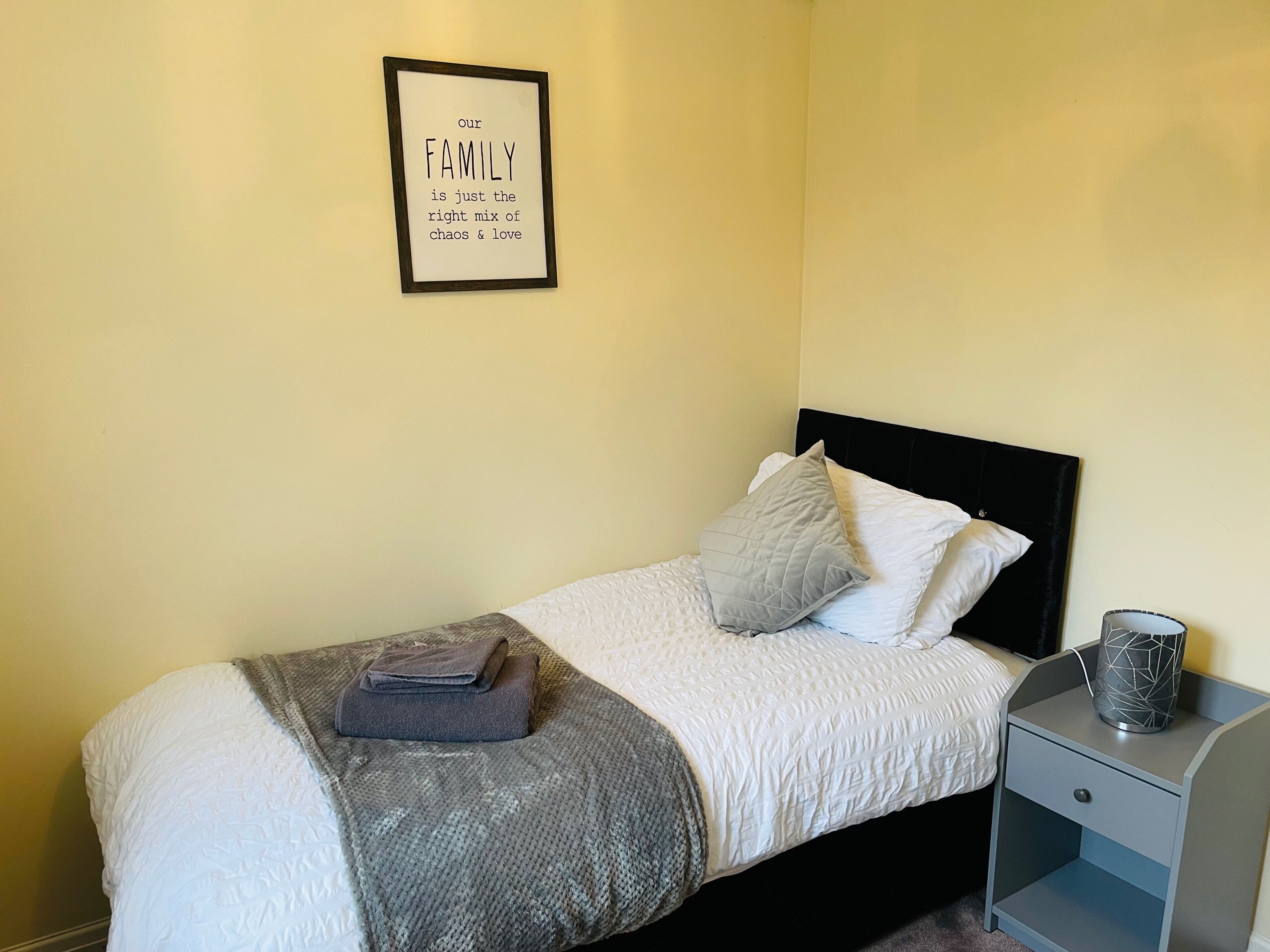 A neatly made bed with a gray blanket and pillows, a bedside table with a lamp, and a framed quote on the wall.