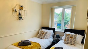 Une chambre d’hôtel avec deux lits, une fenêtre avec des rideaux, une étagère fixée au mur avec des objets décoratifs et une table de chevet avec une lampe.