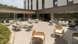 A modern outdoor seating area with wicker furniture and glass walls, overlooking a building with a grid-like facade. - Canvas Pamplona Plazaola  (Pamplona)