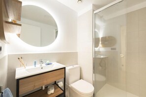 Et moderne badeværelse med et rundt spejl, en vask med toiletartikler, et toilet og en brusekabine med glasvægge.