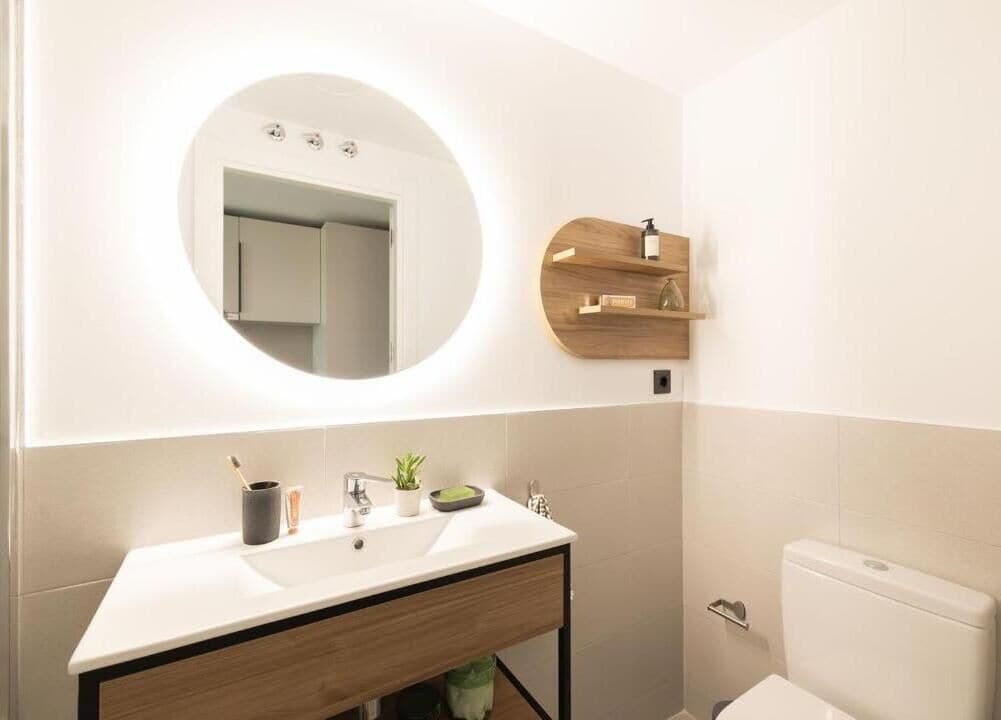 A modern bathroom with a round mirror, two sinks, and a wooden shelf with toiletries.