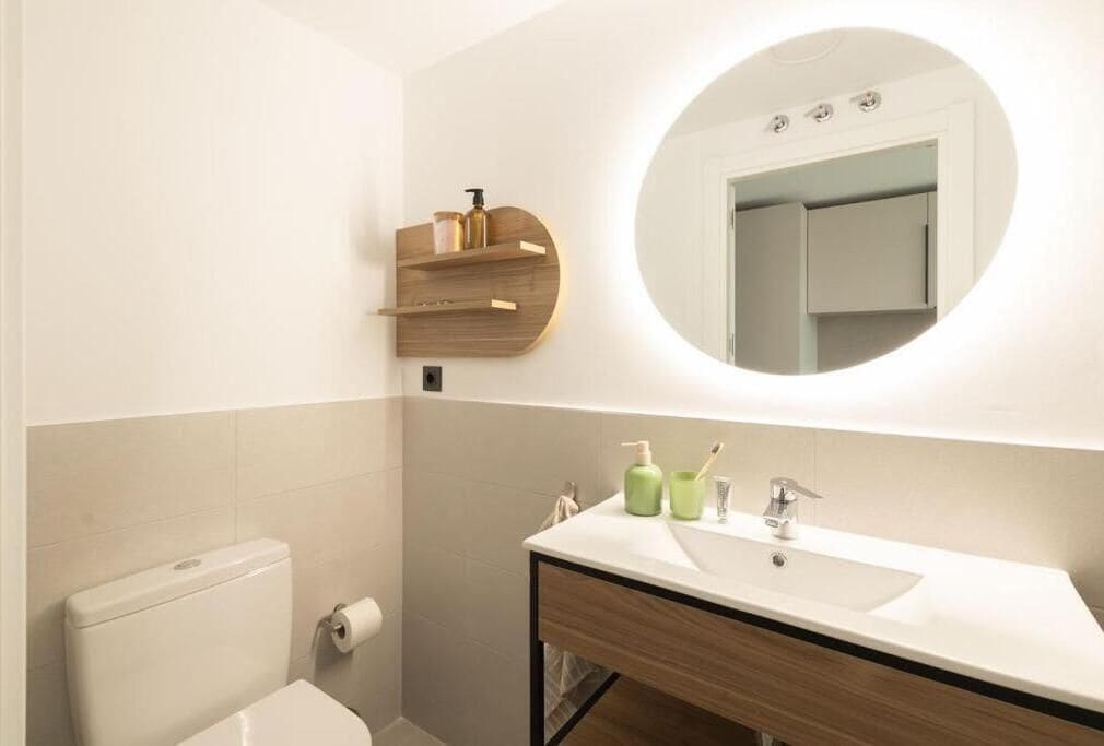 Une salle de bain comprenant des toilettes, un lavabo avec un miroir et une étagère en bois où sont rangés les articles de toilette.
