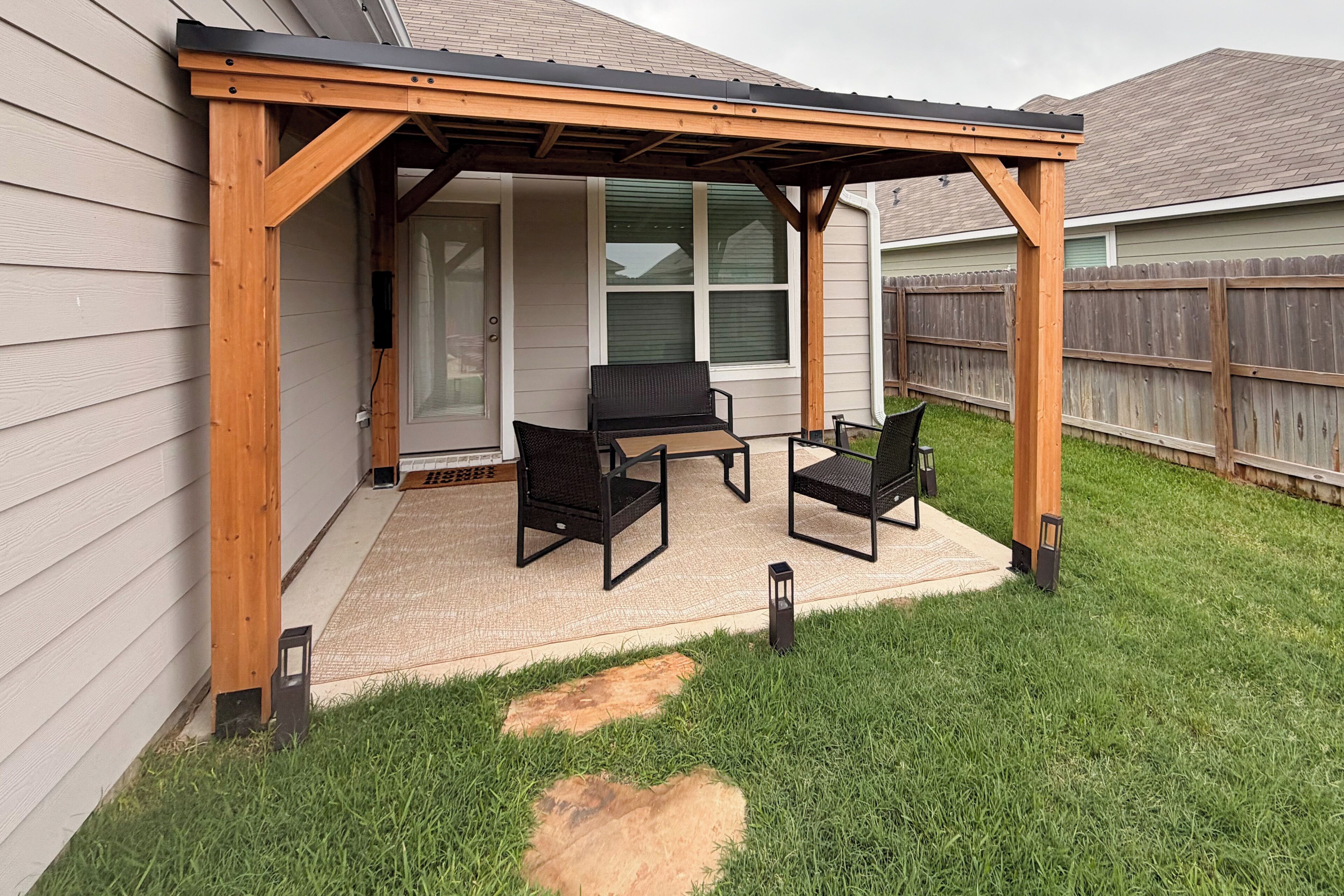 A covered patio with two chairs and a table, a wooden pergola, and a grassy yard.