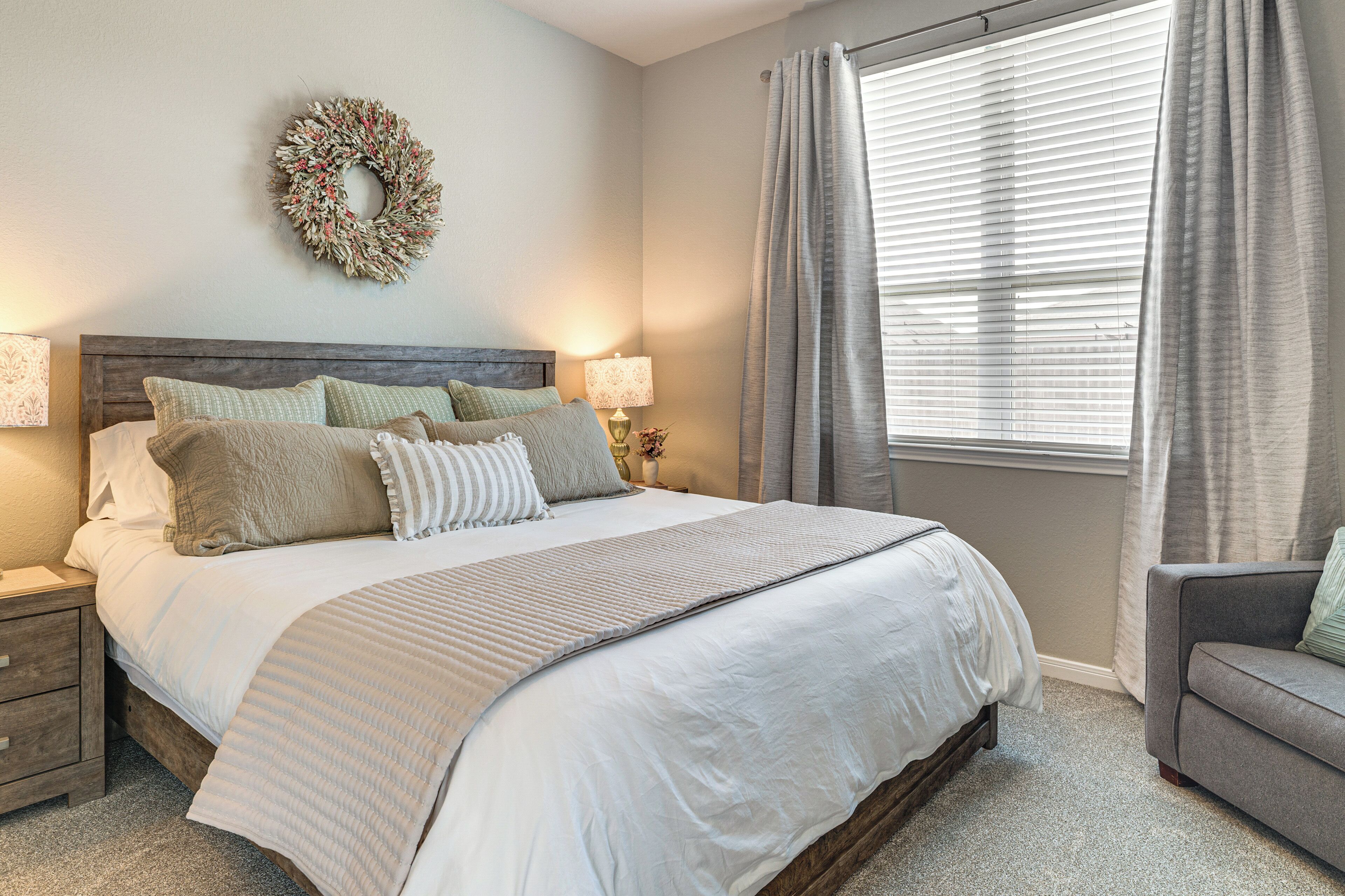 A neatly arranged bedroom with a large bed, a bedside table, a lamp, a grey sofa, and a window with curtains.