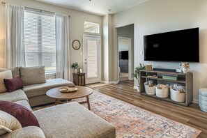 A living room with a sofa, coffee table, and TV. There is a wooden floor, a patterned rug, and a bookshelf with decorative items. - Texas A&M Game Day Hub: Family Retreat w/ Yard (Bryan)