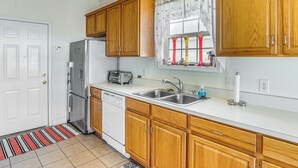 A kitchen with wooden cabinets, a white countertop, a sink, a window with curtains, and a door with a striped mat. - Pool Access & Pond Views: Longs Home Near Golf! (Longs)