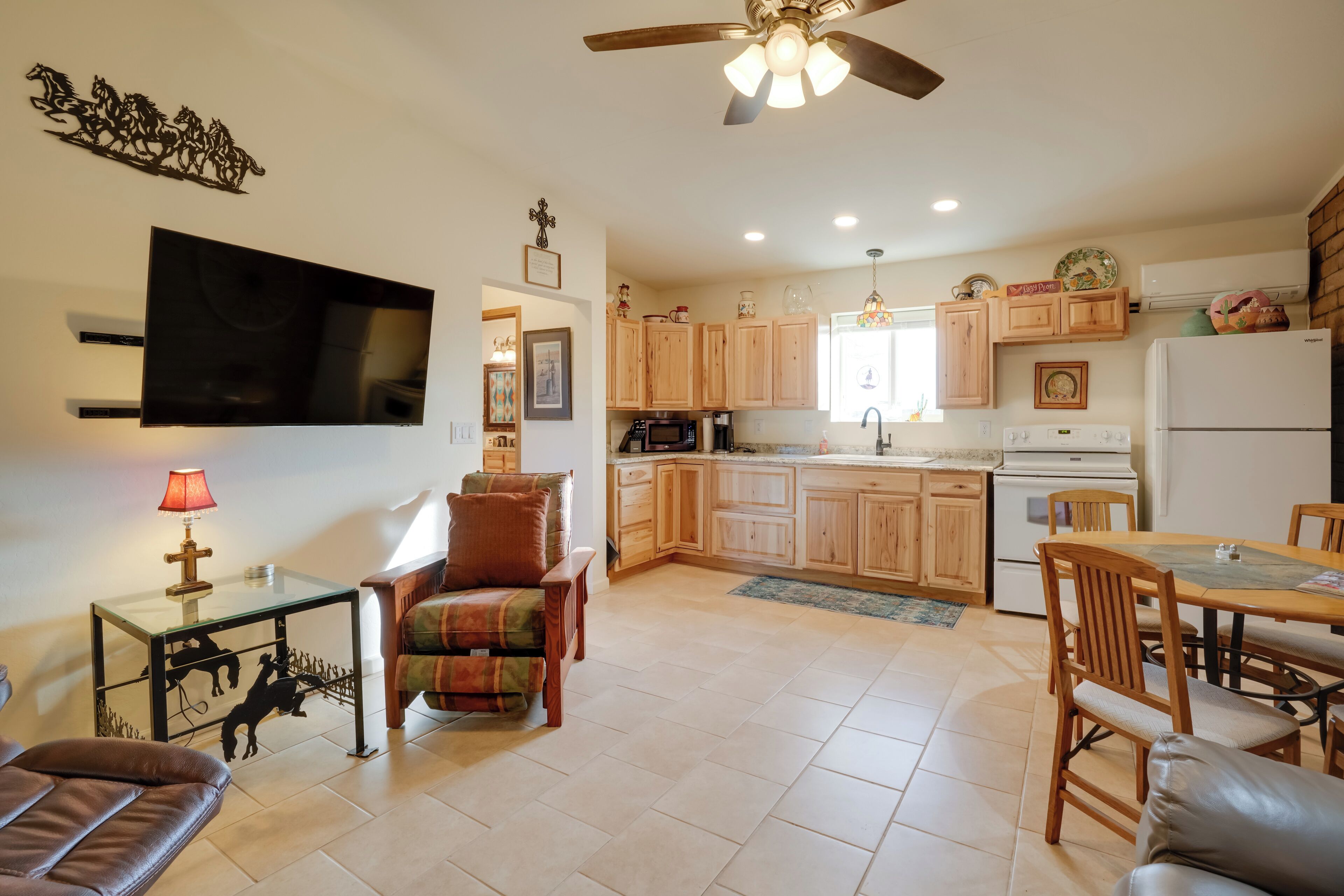 A modern kitchen with wooden cabinets, a dining area with a table and chairs, a flat-screen TV mounted on the wall, and a ceiling fan.