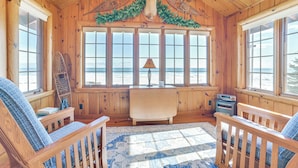 A cozy cabin with wooden walls, a large window with a view of the beach, a fireplace, and a ceiling adorned with antlers. - Waterfront Cottage: Family Getaway at Lake Huron! (East Tawas)